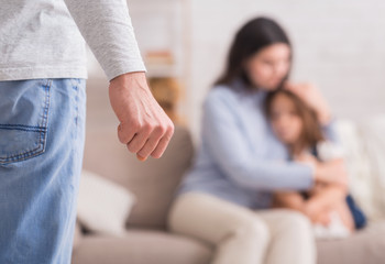 Frau sitzt verängstigt mit Kind in einer Ecke. Der Mann steht vor Ihnen, die Hand zur Faust geballt.