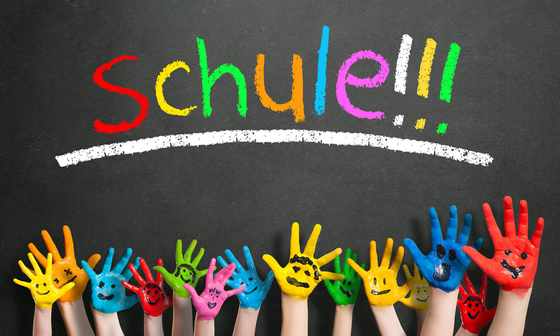 Bunt angemalte Kinderhände, Im Hintergrund eine Tafel, auf der in bunter Schrift "Schule" steht