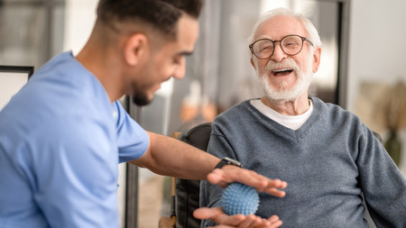 Pfleger spielt mit einem älteren Herr im Rollstuhl Ball