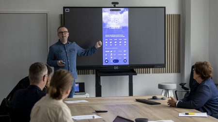 Ein Mann steht vor einem Screen und erläutert den sitzenden Anwesenden am Tisch die Nutzung einer App.