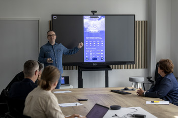 Ein Mann steht vor einem Screen und erläutert den sitzenden Anwesenden am Tisch die Nutzung einer App.